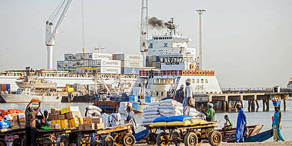 Men loading carts with goods off a ferry and a pirogue in The Gambia.