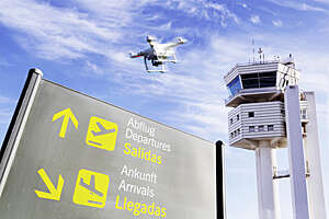 Drone flying low over an airport nearby a control tower, with departures/arrivals panel in the forefront.
