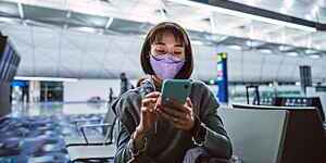 Young woman wearing a protective face mask taps on her smartphone as she waits for a night flight at a quiet airport terminal gate.